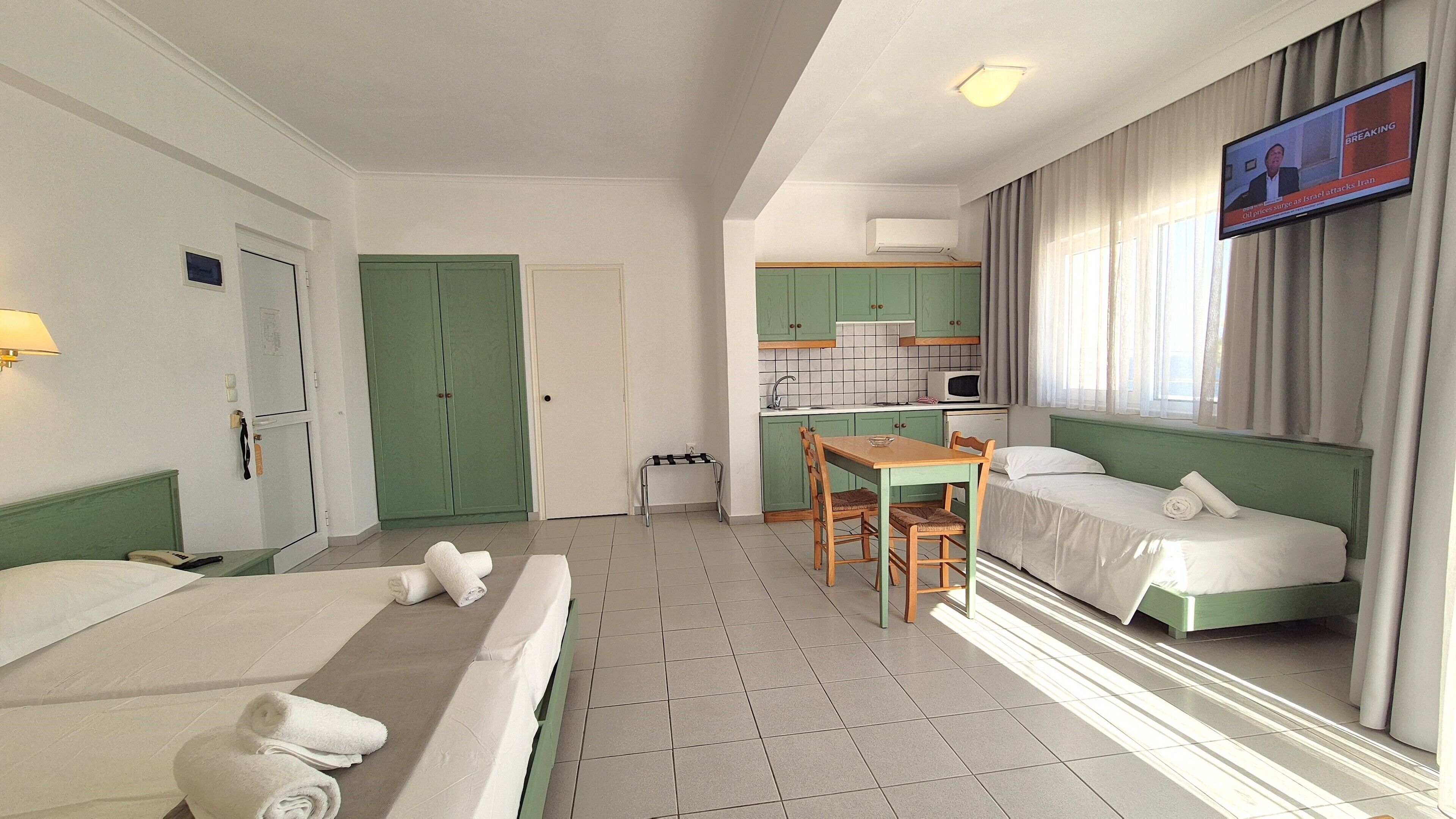 A hotel room with two beds, a small kitchenette, a dining table, and a TV on the wall.