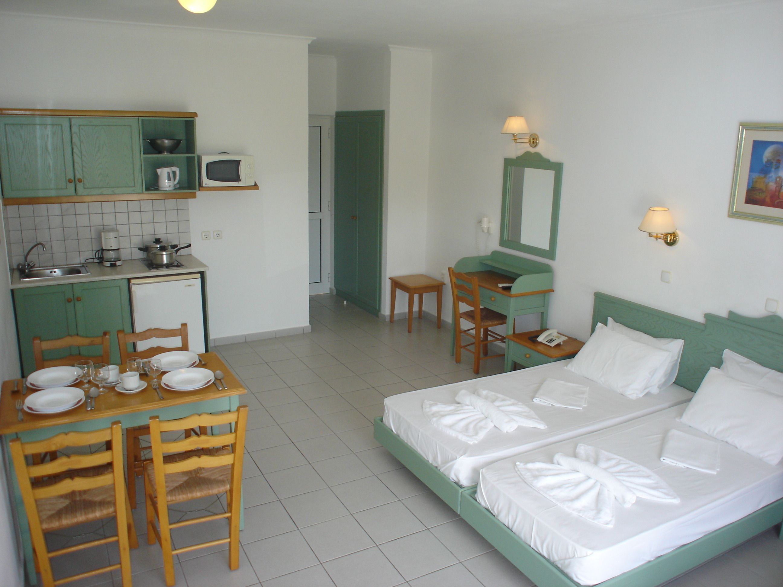 A hotel room with a bed, a desk, a chair, a table set for dining, and a kitchenette.