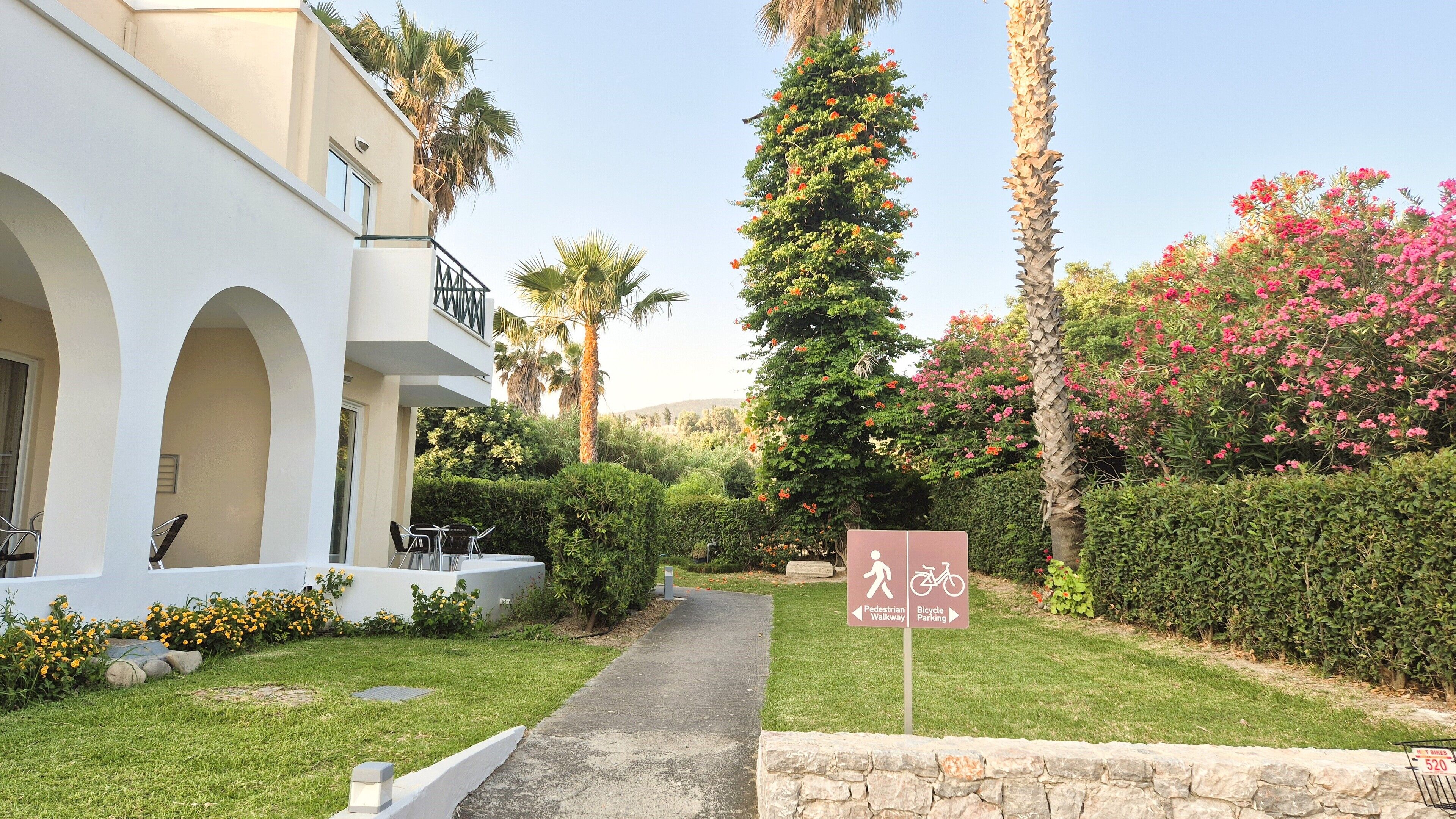 A pathway leading to a building with a balcony, surrounded by greenery and a sign indicating pedestrian and bicycle parking.