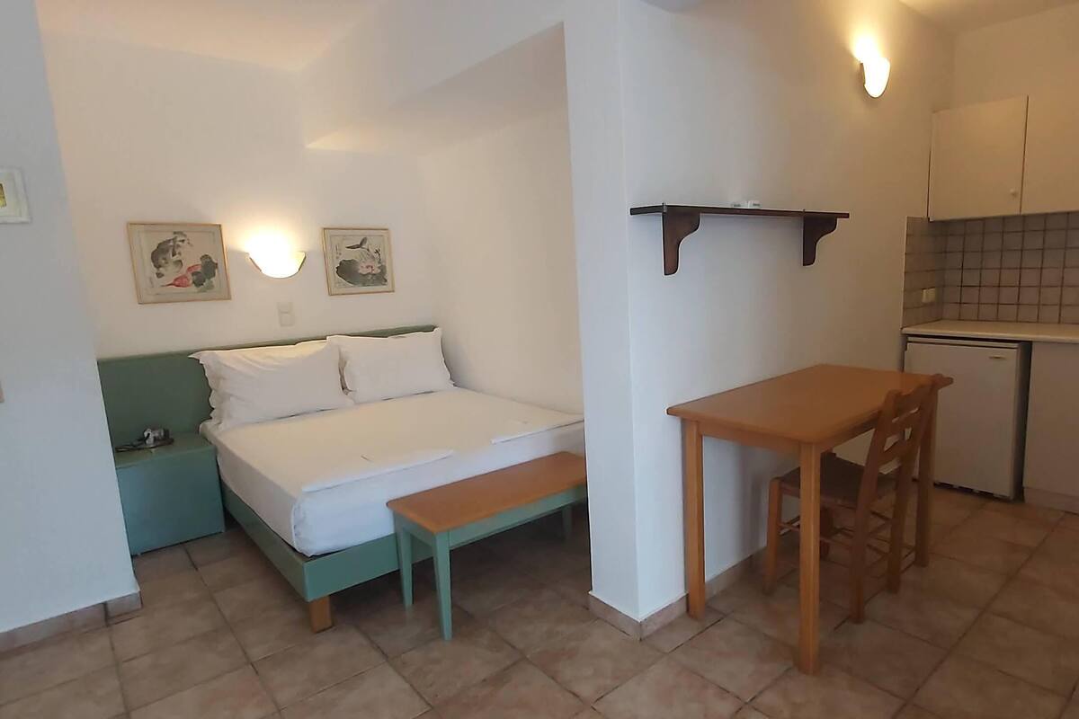 Ein kleines Zimmer mit einem Bett, einem Tisch, einer Sitzbank, einer Wandablage und einer Küchenzeile.