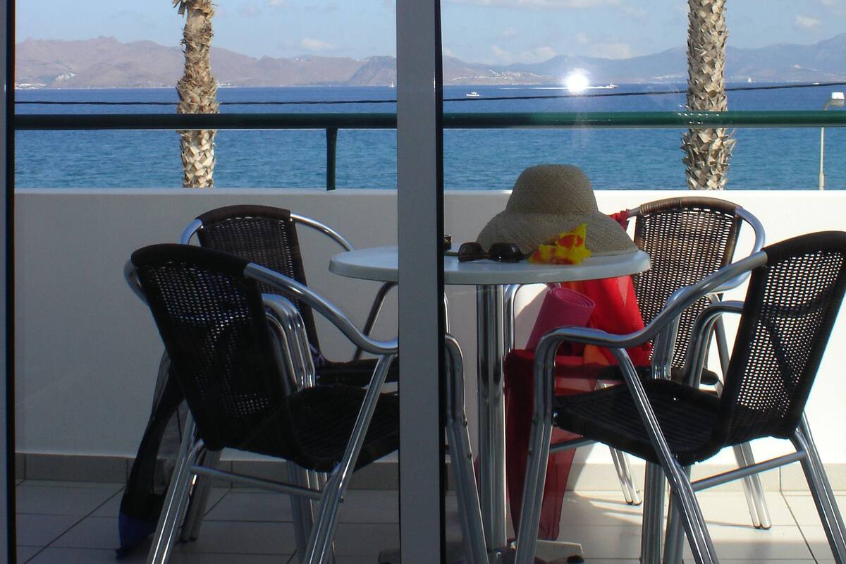 Ein Balkon mit zwei Palmen, einem Tisch mit einem Hut und Stühlen.