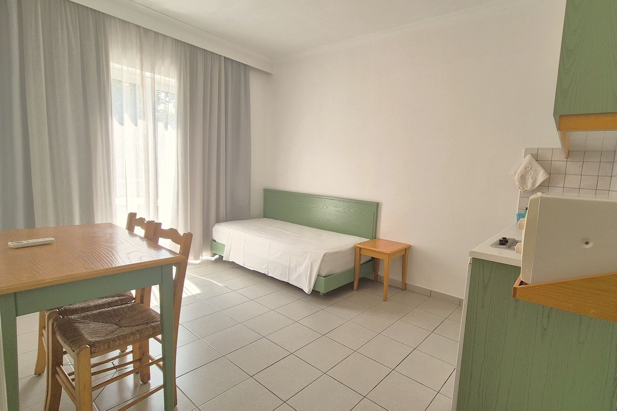 A small, clean room with a bed, a table, a chair, and a kitchenette.