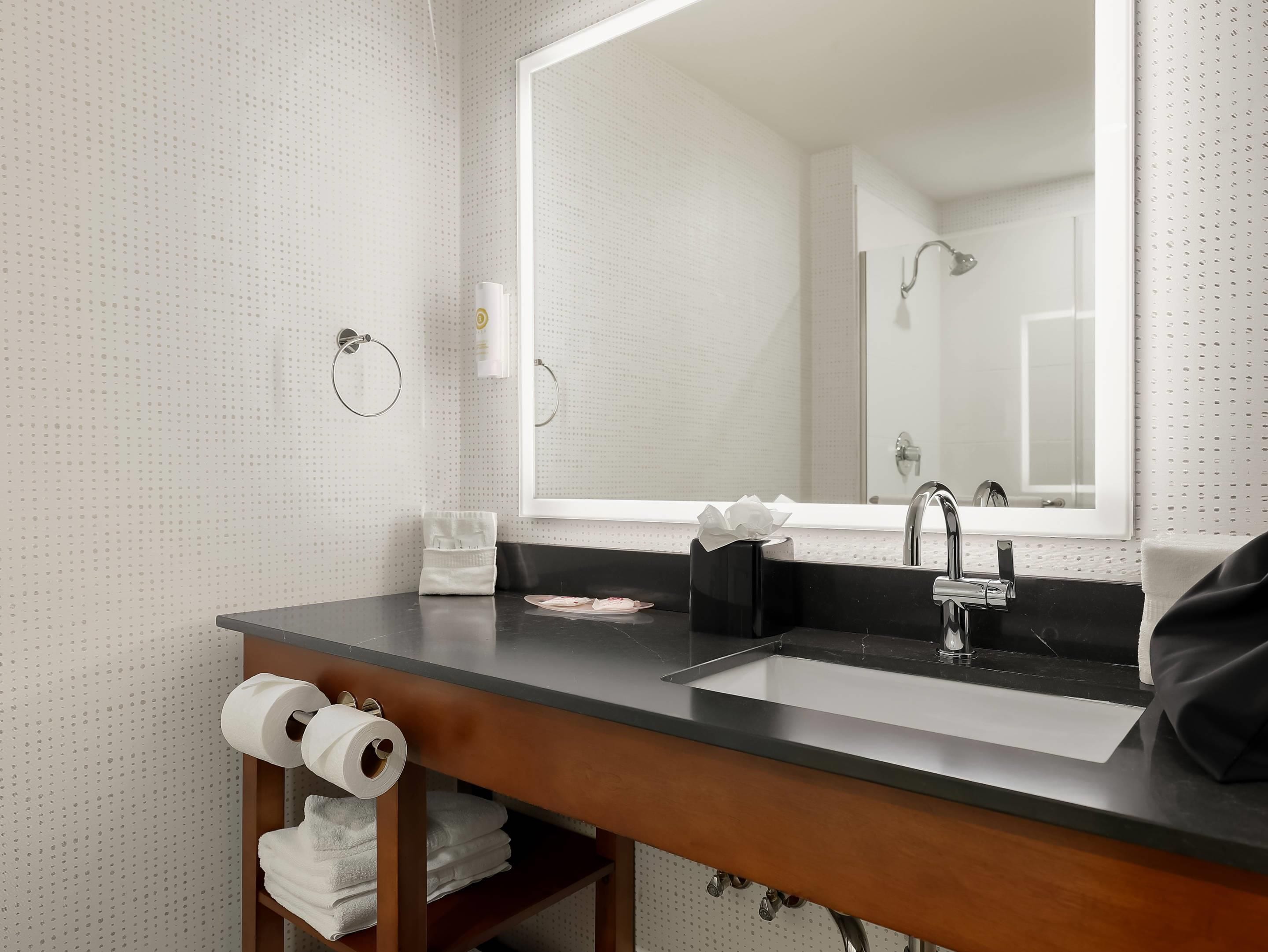 Un baño con un espejo grande, dos lavamanos, un perchero para toallas y un estante con toallas y papel higiénico.