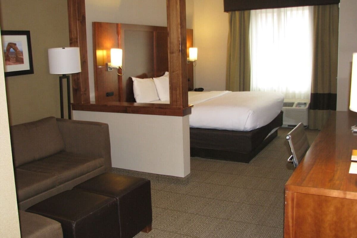 Una habitación de hotel con cama, sofá, escritorio, espejo y ventana con cortinas.