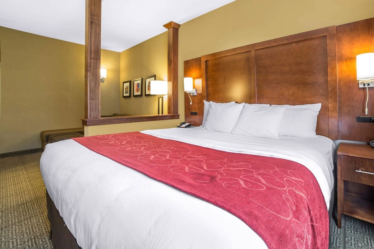 Una habitación de hotel con una cama grande, un cabecero de madera, una manta roja y una mesita de noche con una lámpara.