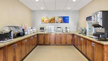 Un área de desayuno con una máquina de café, una exhibición de pan y un dispensador de jugo.