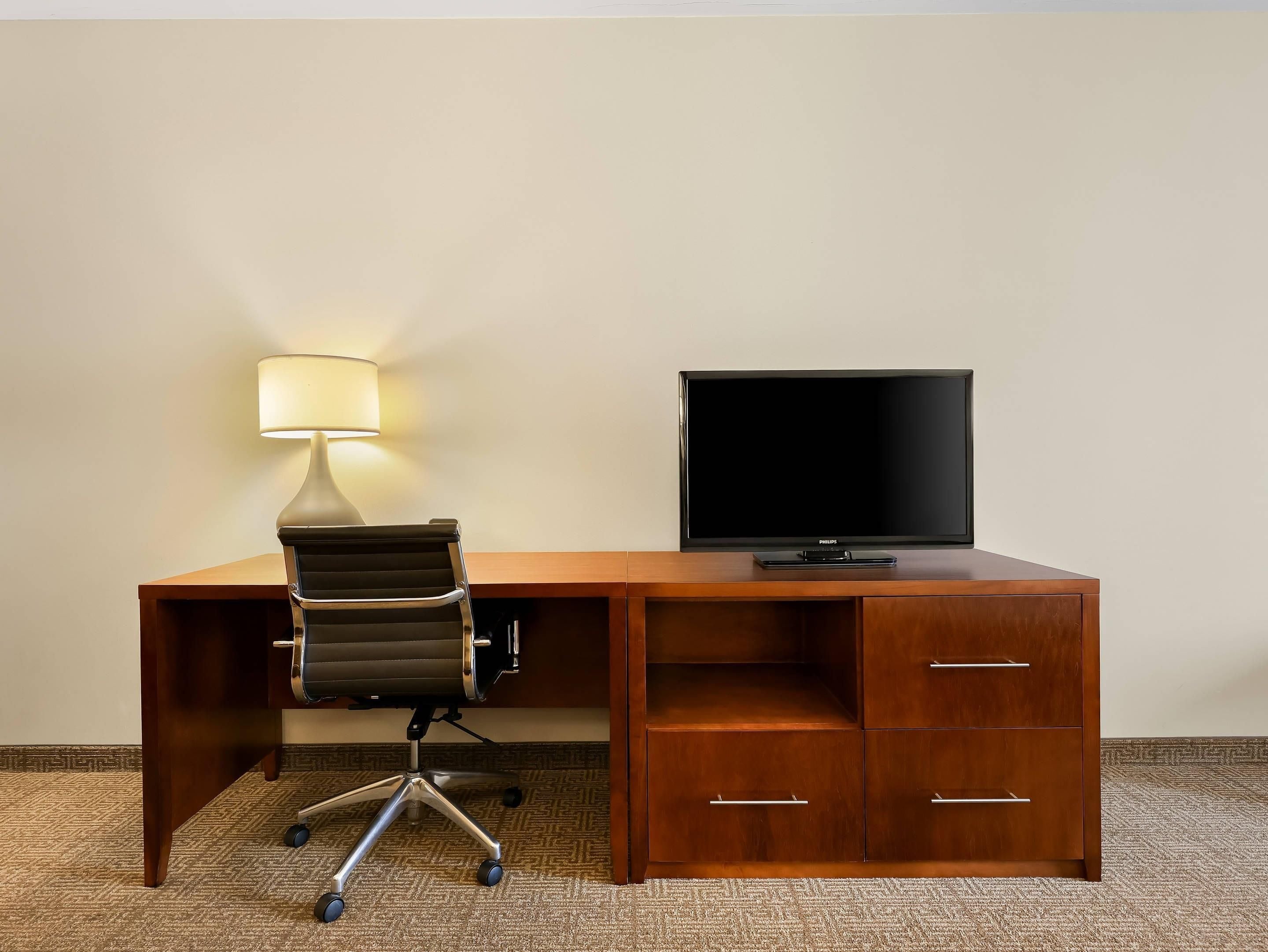 A hotel room with a wooden desk, a black office chair, a lamp, and a television.