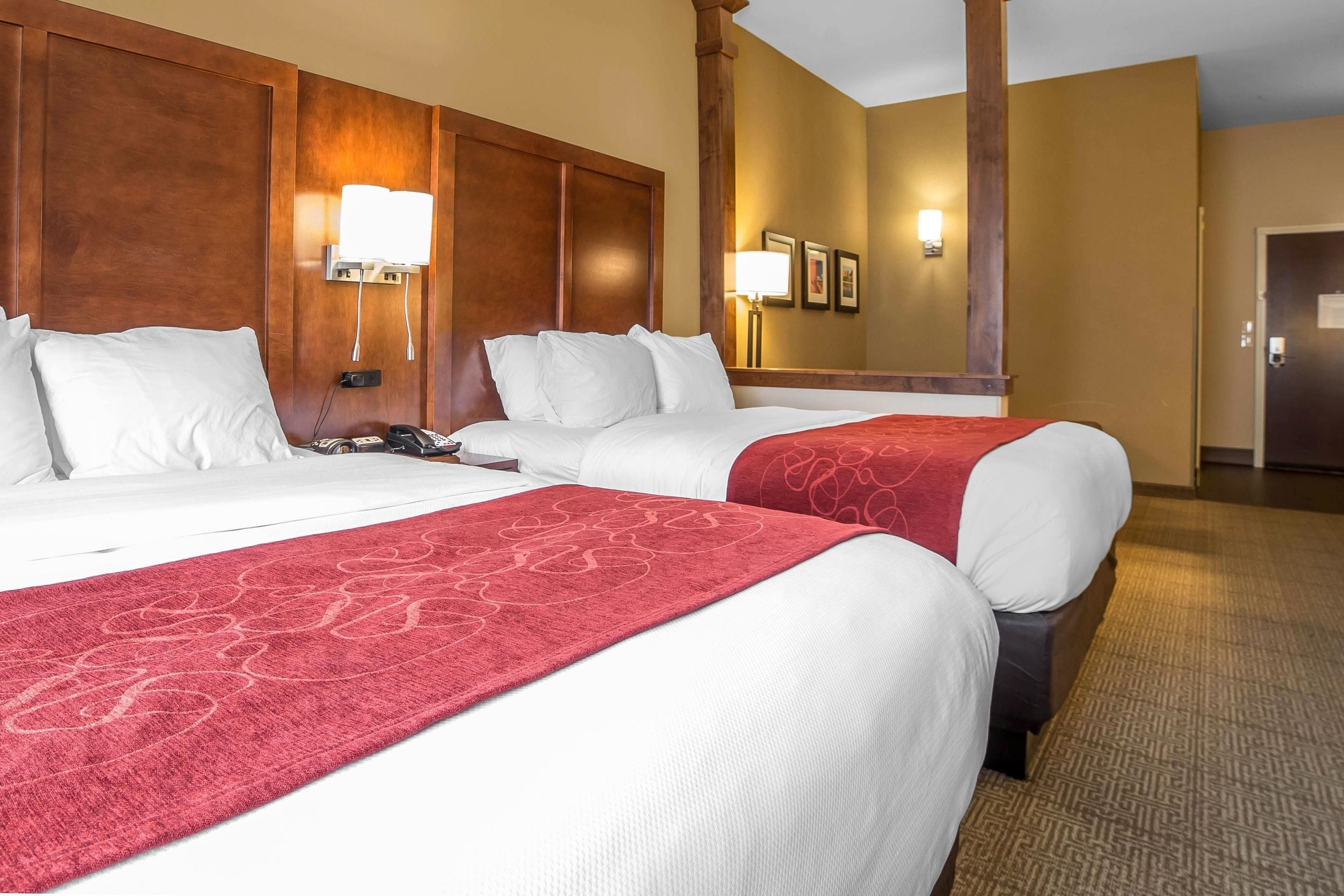A hotel room with two beds, a wooden headboard, a red patterned runner, and a wall-mounted lamp.