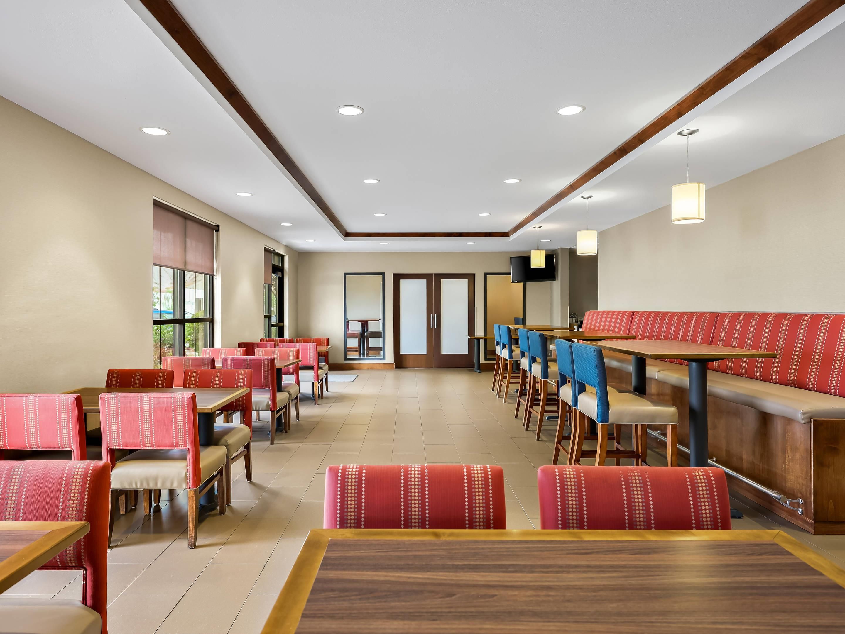 A hotel dining area with multiple tables and chairs, a long bench, and a television mounted on the wall.