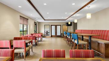 A hotel dining area with multiple tables and chairs, a long bench, and a television mounted on the wall.