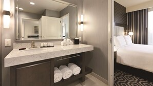 Une chambre d’hôtel dotée d’un meuble-lavabo en marbre, d’un grand miroir, d’un lit avec du linge de maison blanc et d’un tapis à motifs.