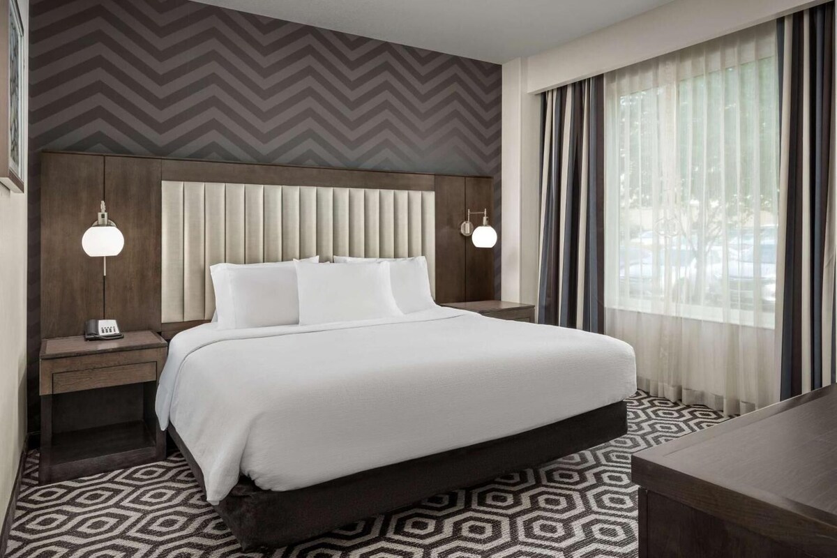 A hotel room with a bed, bedside tables, a patterned carpet, a chevron-patterned wall, and a window with curtains.