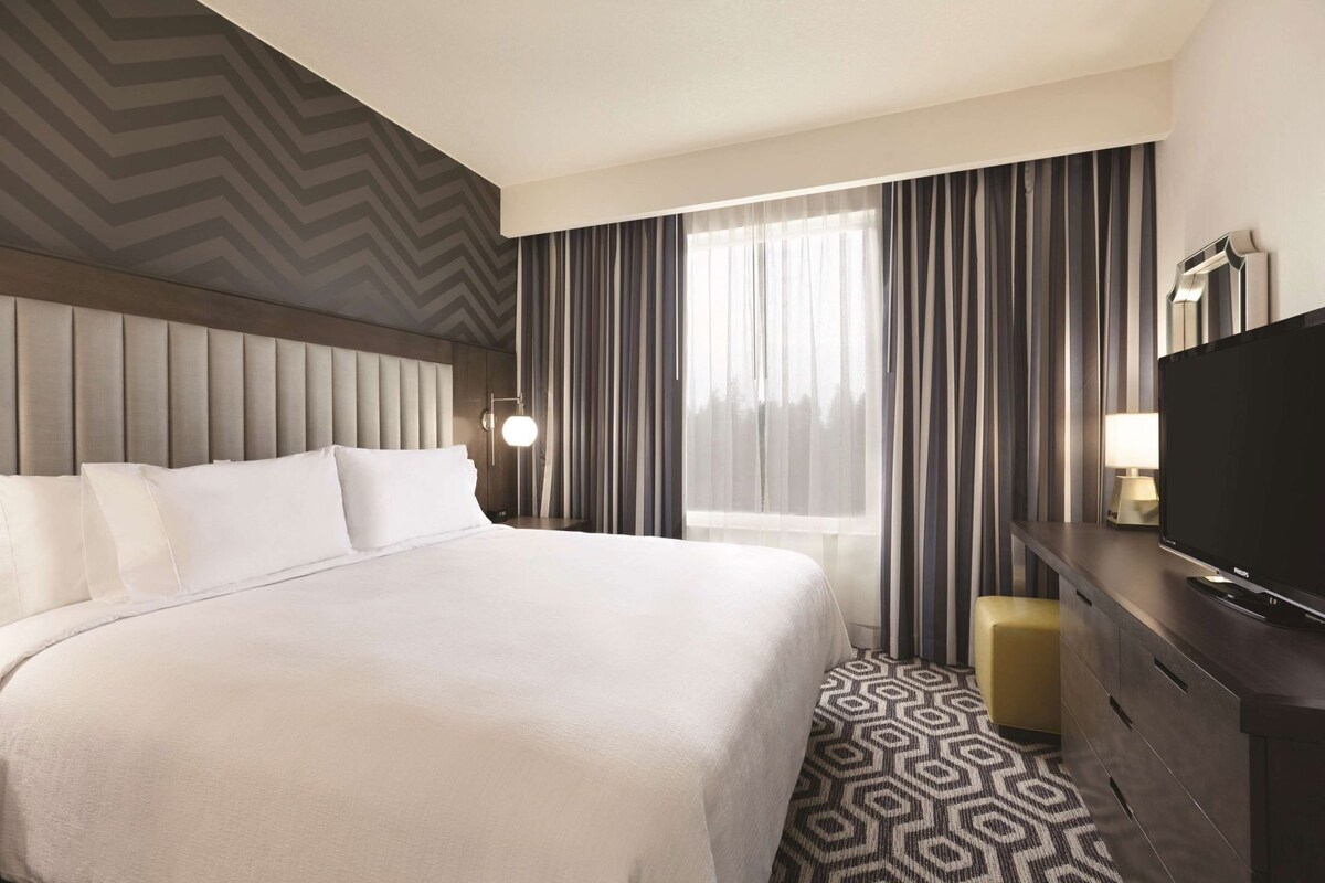 A hotel room with a large bed, a television, a desk with a lamp, and a window with curtains.