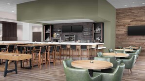 Un espace bar moderne avec des tables en bois et des chaises vertes, un téléviseur à écran plat fixé au mur, et des étagères remplies de bouteilles et de verres.