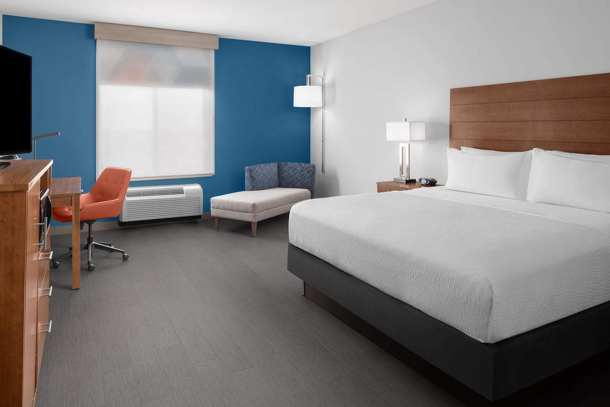 Une chambre d’hôtel avec un grand lit, un bureau avec une chaise, un canapé, une télévision et une fenêtre avec des stores.