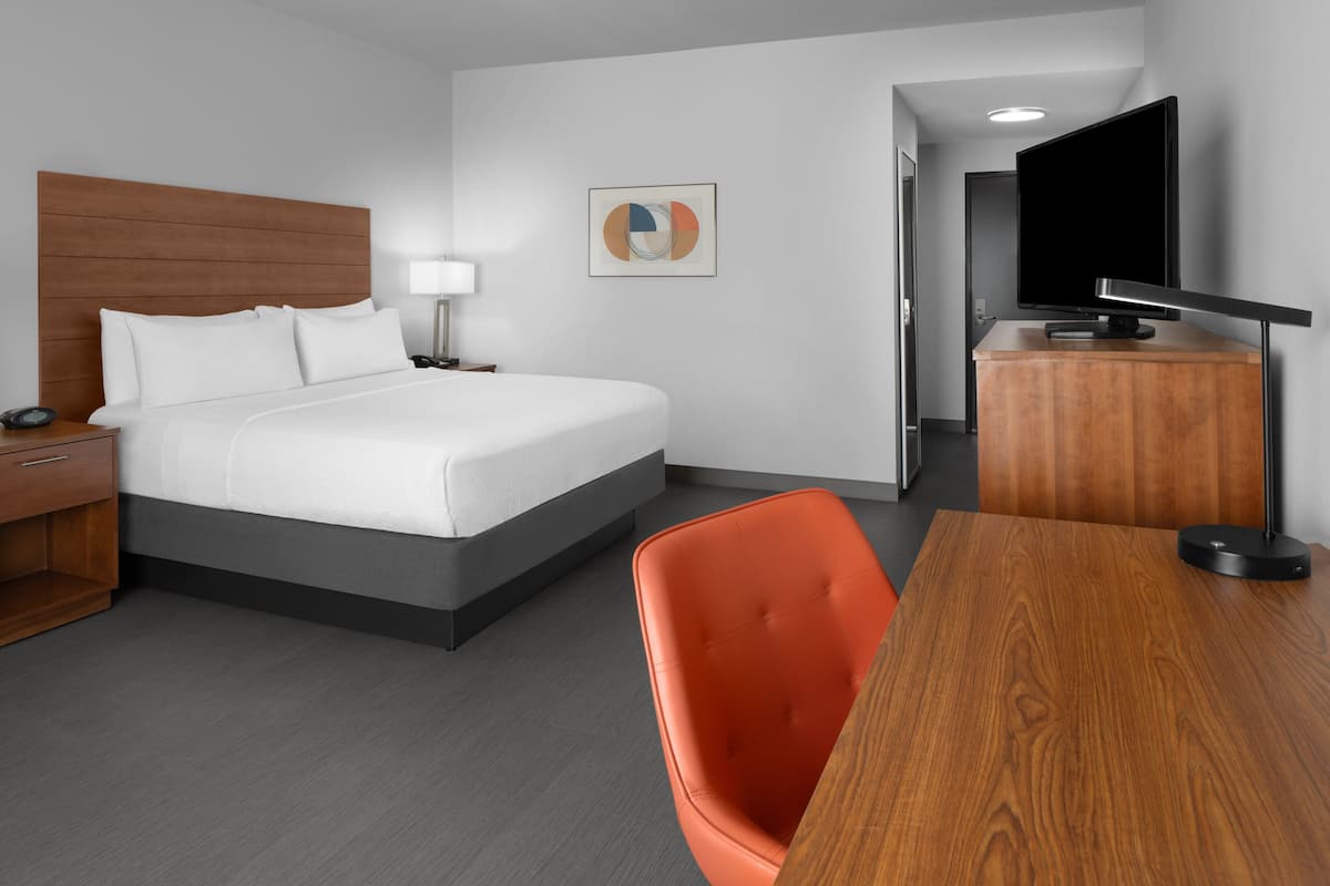 Une chambre d’hôtel moderne, équipée d’un lit, d’un bureau, d’une chaise, d’une télévision et ornée d’un tableau au mur.