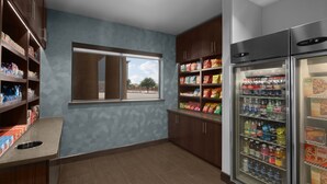 A kitchenette with a window, shelves stocked with snacks, a refrigerator with beverages, and a trash bin. - Holiday Inn Express & Suites Midland South I-20 by IHG (Midland)