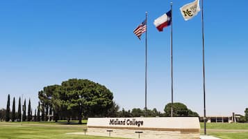 A grassy area with a sign for Midland College, three flagpoles with flags, and a large tree in the background.