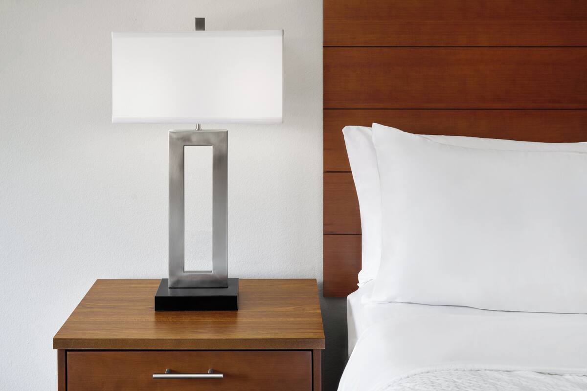 Une table de chevet en bois, surmontée d’une lampe moderne, avec un tiroir, et un oreiller blanc sur un lit.