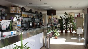 A modern bar area with a stainless steel counter, various bottles, and a trash bin. There are potted plants and a seating area with white chairs. - Hotel Cliché (San Lucido)
