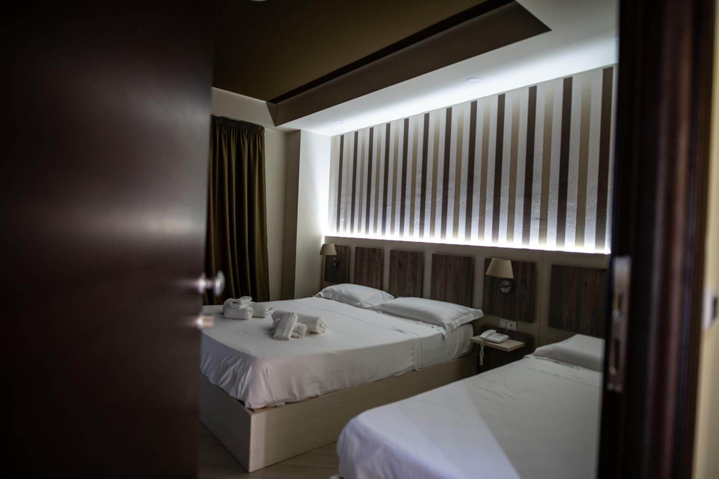 Habitación de hotel con dos camas, un ventanal grande con persianas verticales, una mesita de noche con lámpara y una puerta entreabierta.