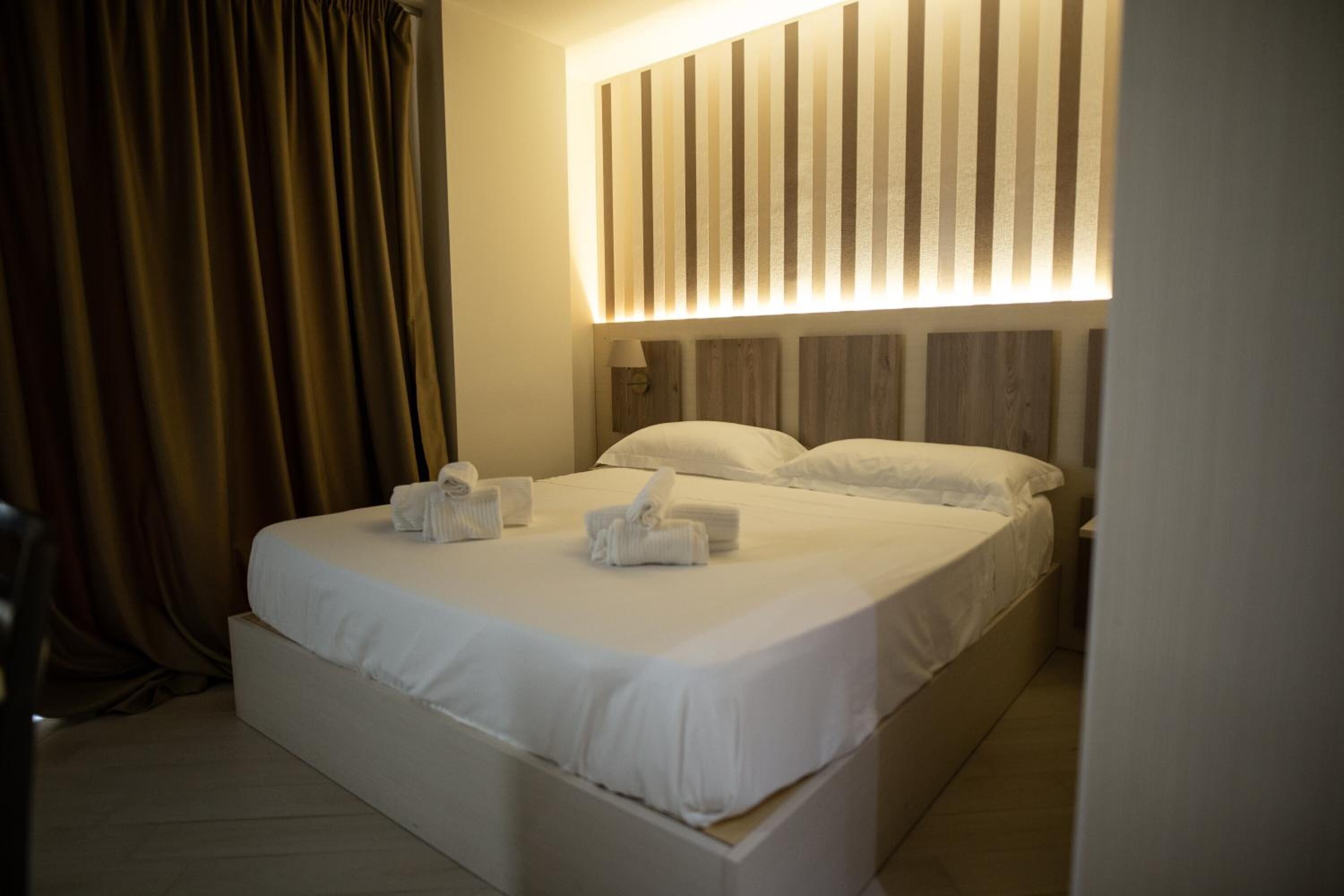 A hotel room with a large bed, a headboard with vertical slats, and a curtain on the left.