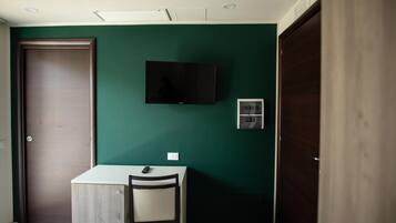 Una habitación con pared verde oscuro, un televisor montado, una mesita con una silla y una puerta de madera.