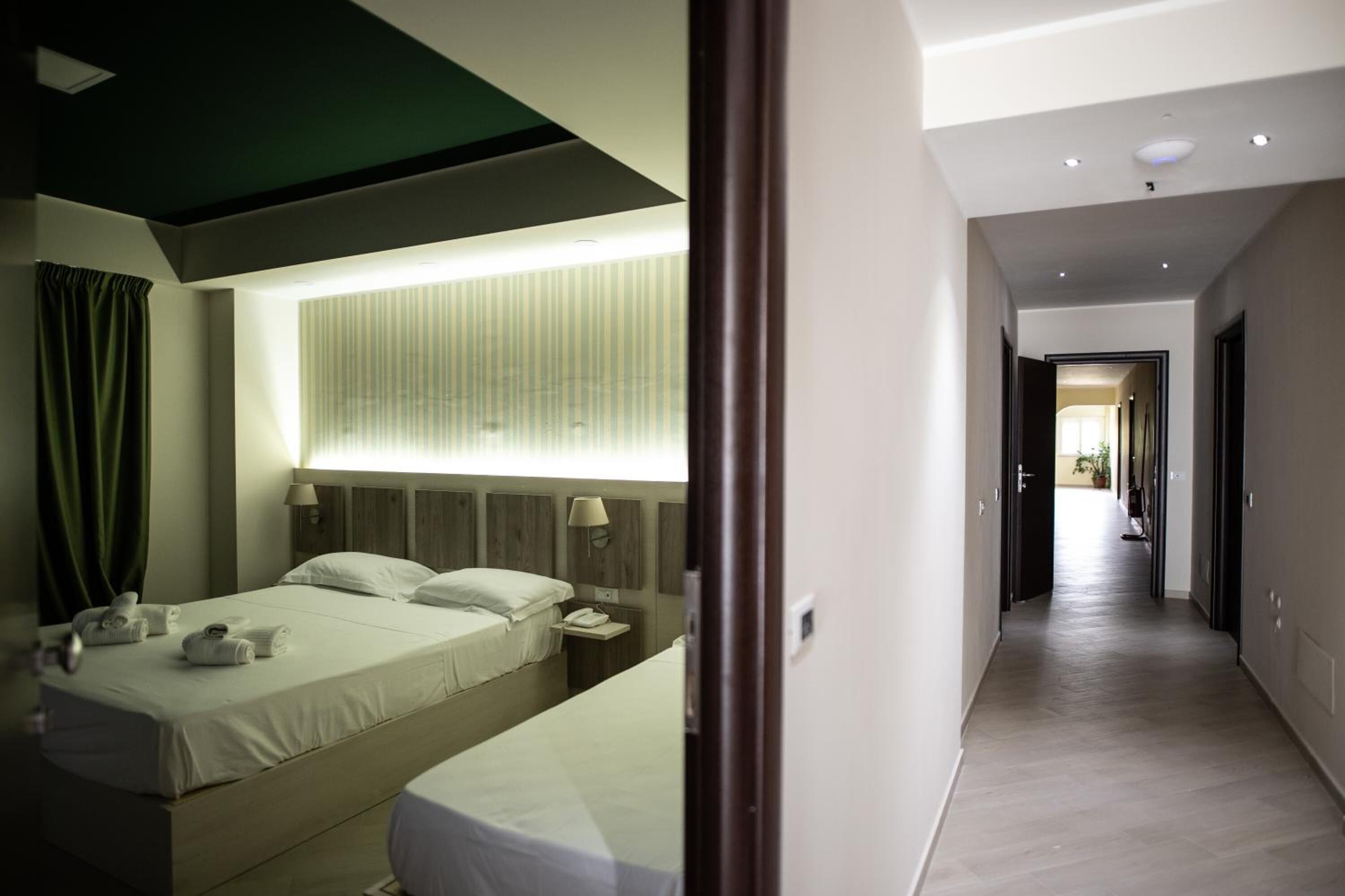 A hotel room with a bed, bedside table, lamp, and a view of a corridor.