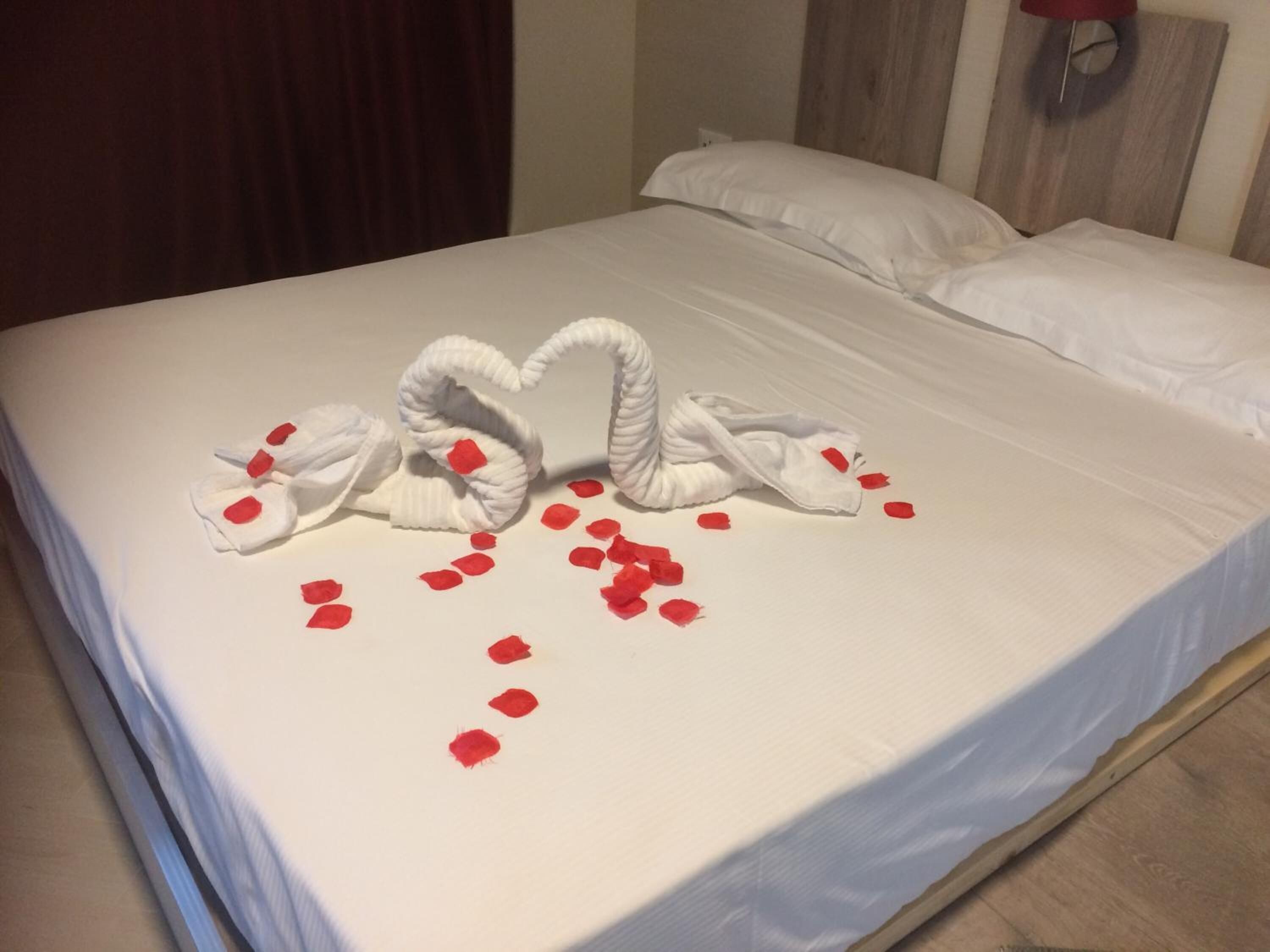 A bed with white linens and a decorative swan made of towels on the bedspread, with red rose petals scattered around.