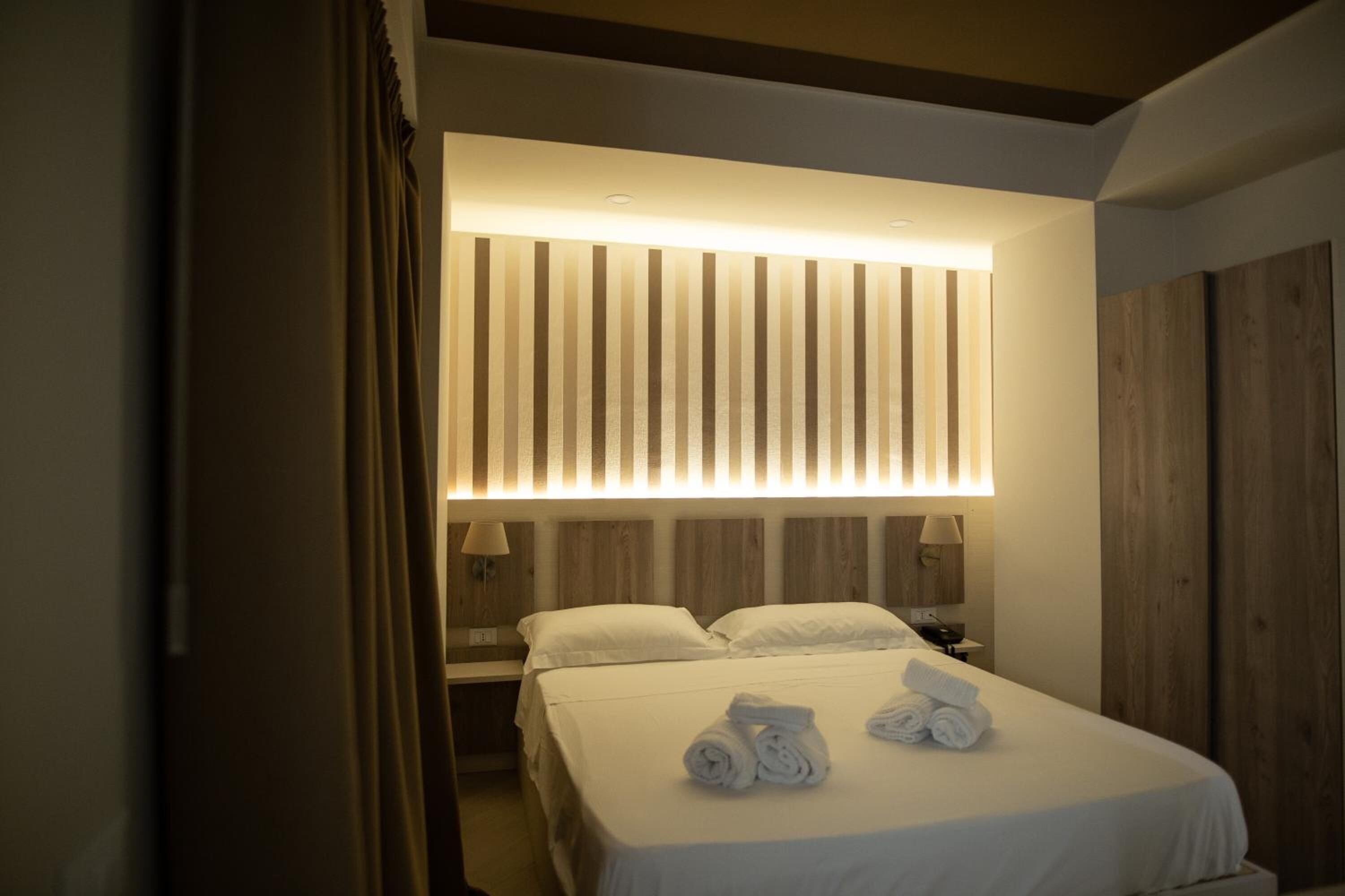 A hotel room with a large bed, two bedside tables, a headboard with vertical slats, and a wall-mounted lamp.