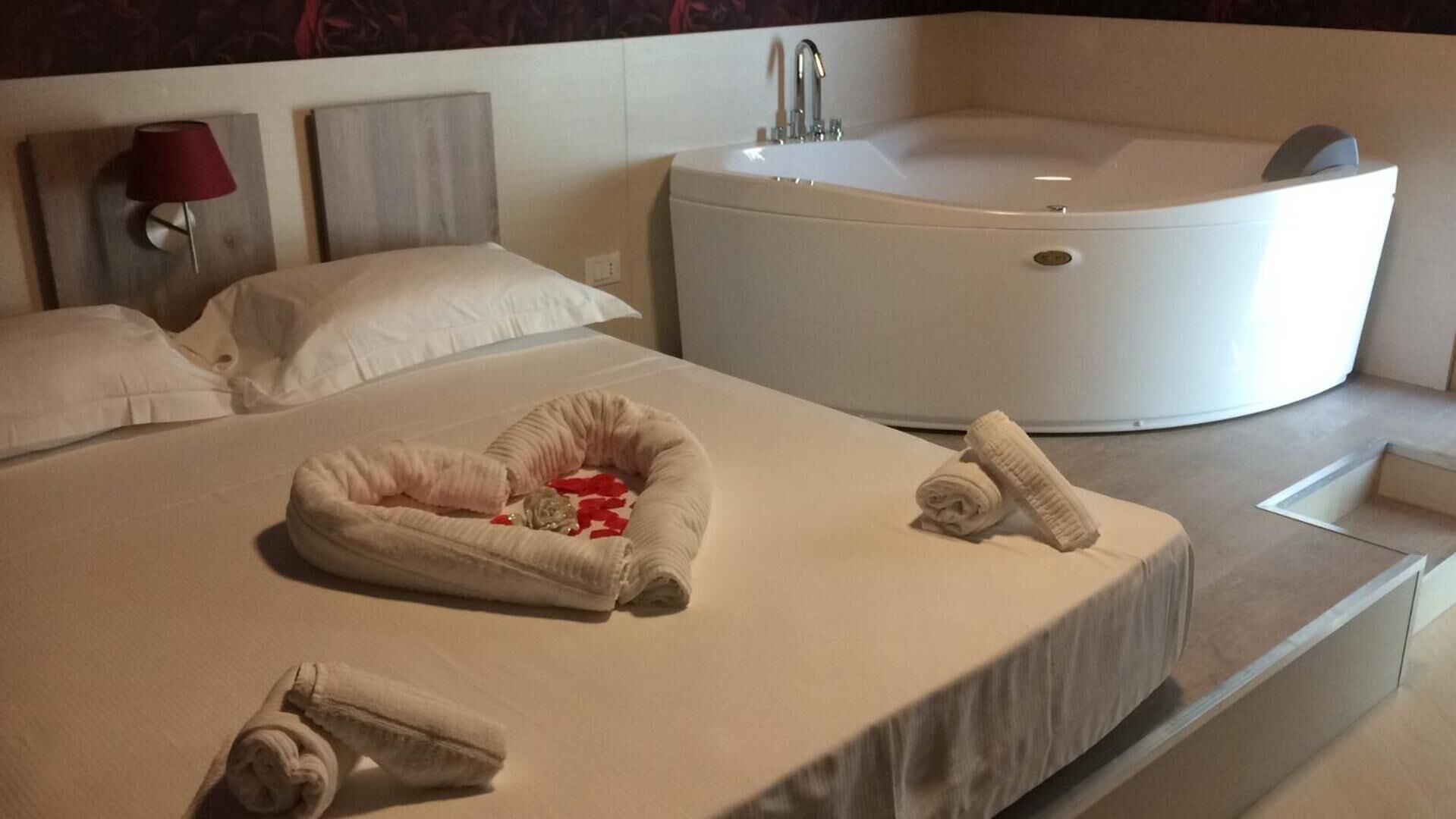 Une chambre d’hôtel moderne avec une grande baignoire, un lit bien fait agrémenté d’un tapis en forme de cœur, et une lampe rouge sur la table de chevet.
