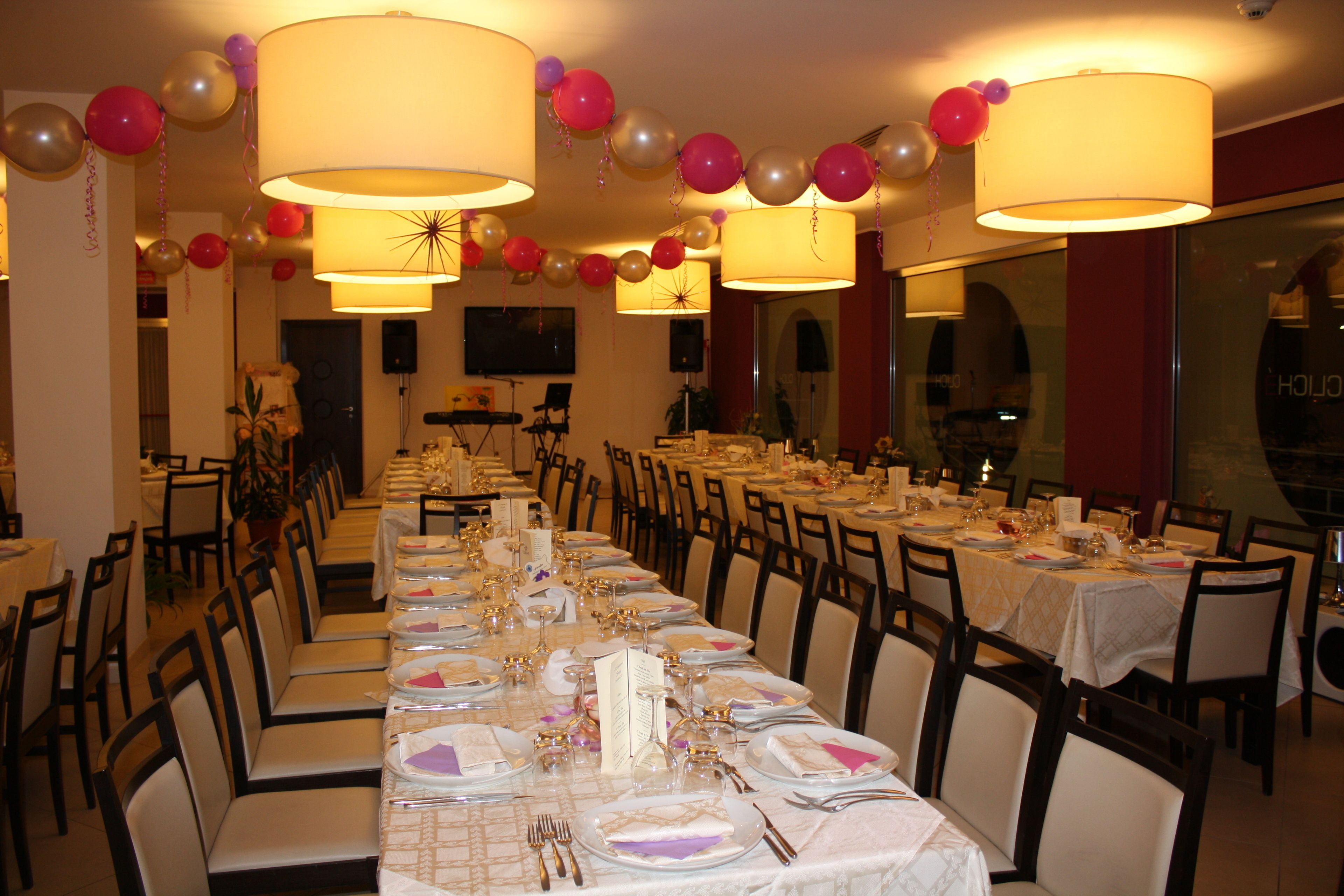 A banquet hall with long tables set for dining, chairs arranged around them, and balloons decorating the ceiling.