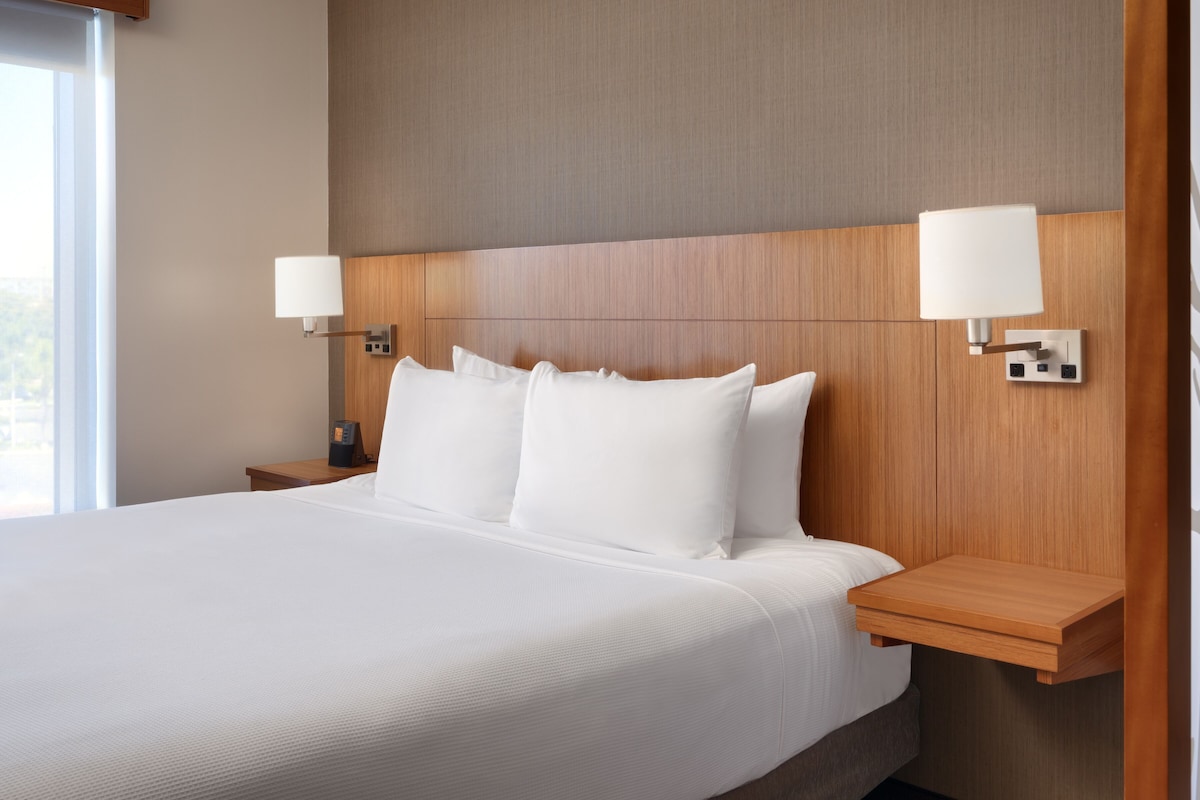A hotel room with a large bed, two bedside tables, a nightstand, and a wall-mounted lamp.