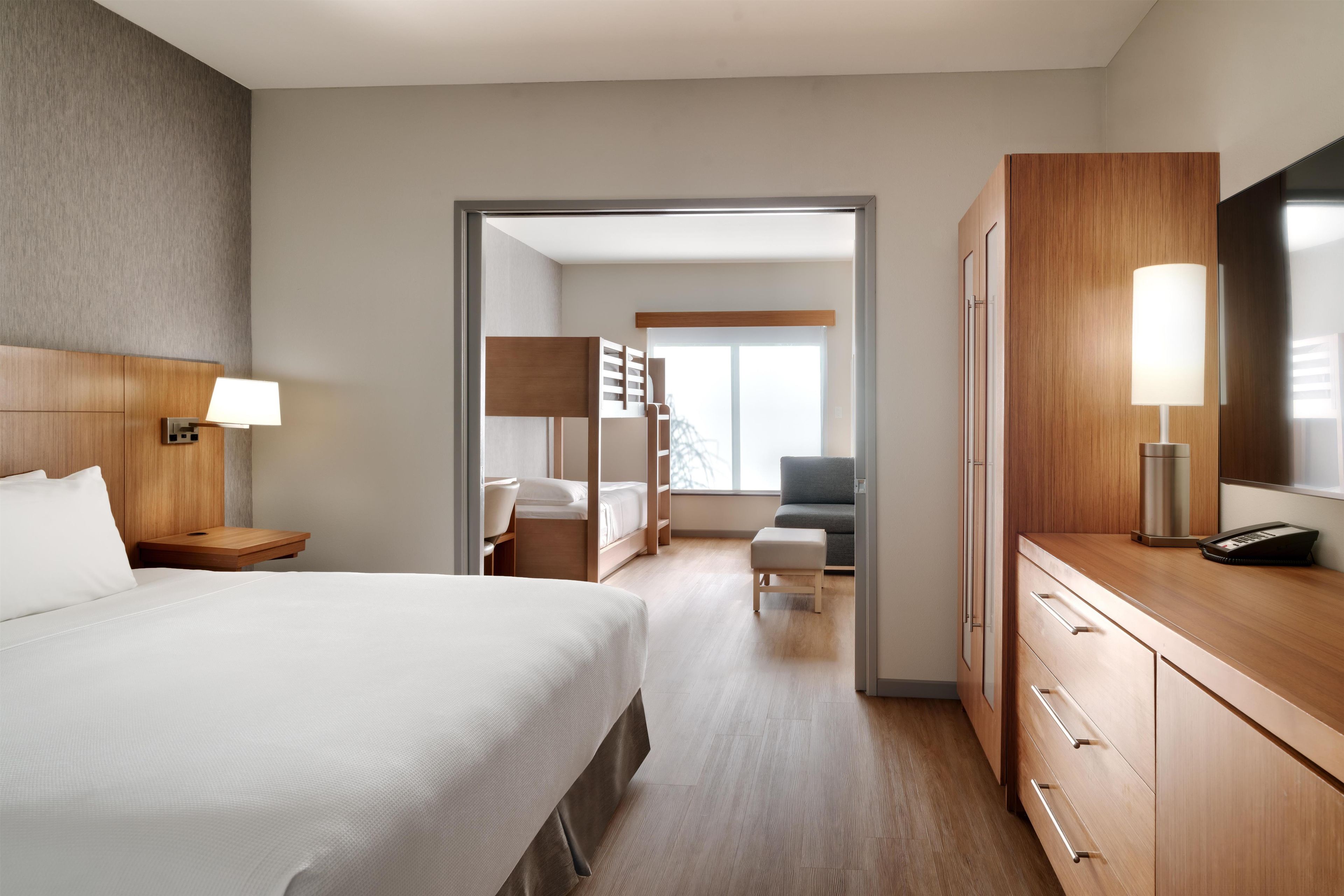 Habitación de hotel con cama, mesita de noche, lámpara, armario y vistas a una zona de estar a través de una puerta abierta.