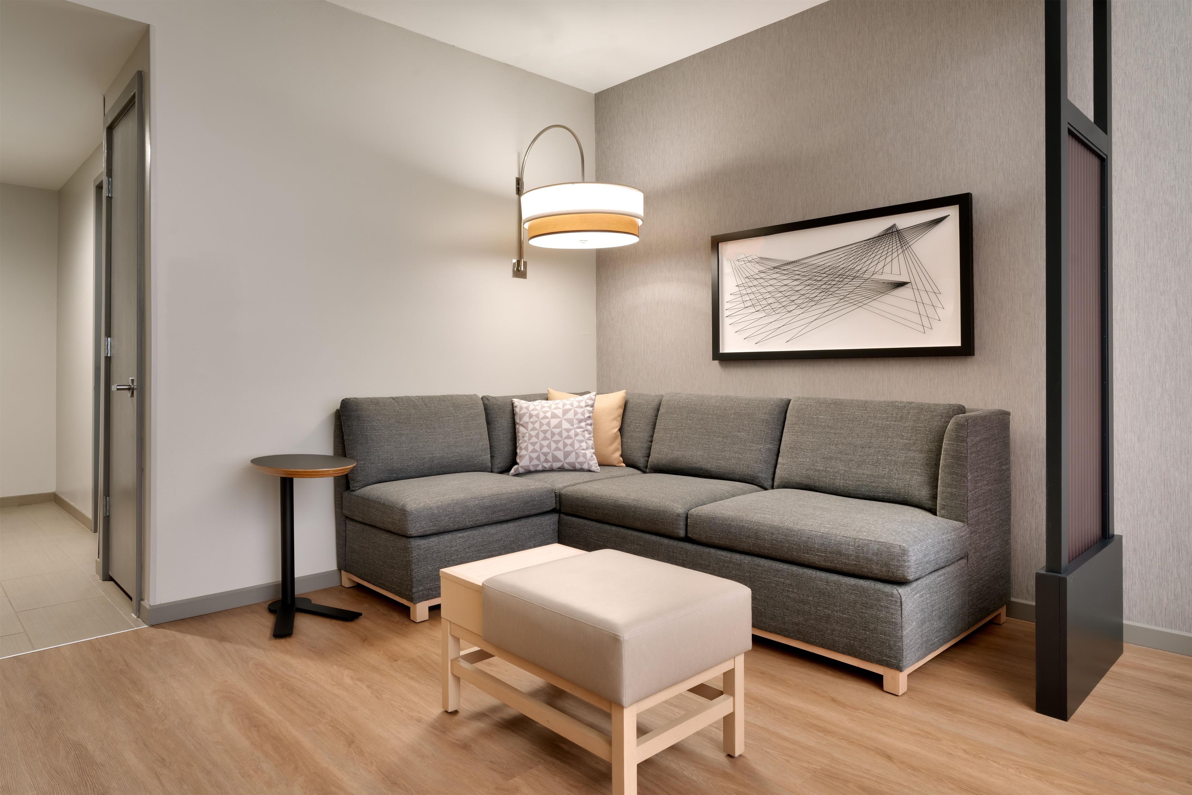 Ein modernes Wohnzimmer mit einem grauen Ecksofa, einem kleinen runden Beistelltisch, einer Bank, einer Stehlampe und einem gerahmten abstrakten Kunstwerk an der Wand.