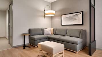 Ein modernes Wohnzimmer mit einem grauen Ecksofa, einem kleinen runden Beistelltisch, einer Bank, einer Stehlampe und einem gerahmten abstrakten Kunstwerk an der Wand.