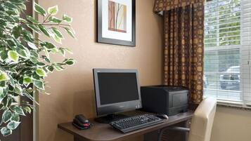 A computer desk with a monitor, keyboard, and printer. There is a framed picture on the wall and a plant to the left.