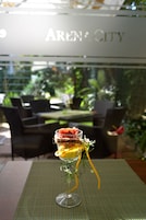 Un postre en un vaso sobre una mesa con un mantel individual verde, con una vista de un restaurante llamado "Aren City" al fondo.