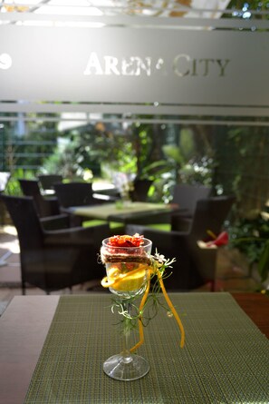 Sobremesa em copo sobre uma mesa com um jogo americano verde, com vista para um restaurante chamado "Aren City" ao fundo.