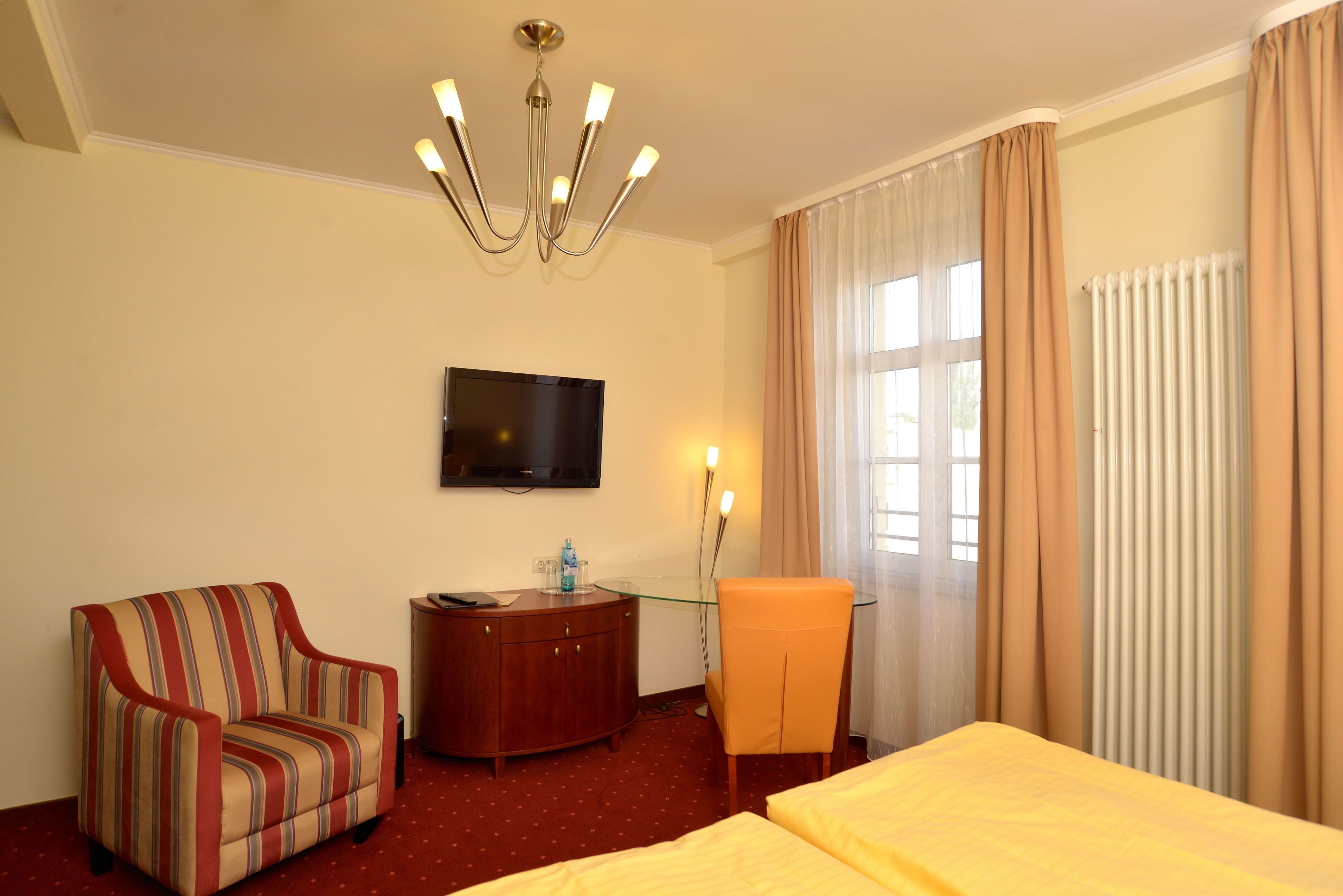 Habitación de hotel con televisor montado en la pared, un sillón a rayas, cómoda de madera, mesa de vidrio, una silla naranja y una cama con colcha amarilla.