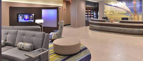 A lobby with a sofa, armchair, and ottoman, a reception desk, a television screen, and a large wall mural.