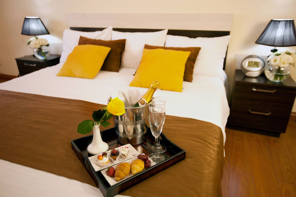 Ein ordentlich bezogenes Bett mit einem Frühstücksbrett, das Croissants, Obst und eine Champagnerflasche enthält, in einem Hotelzimmer.