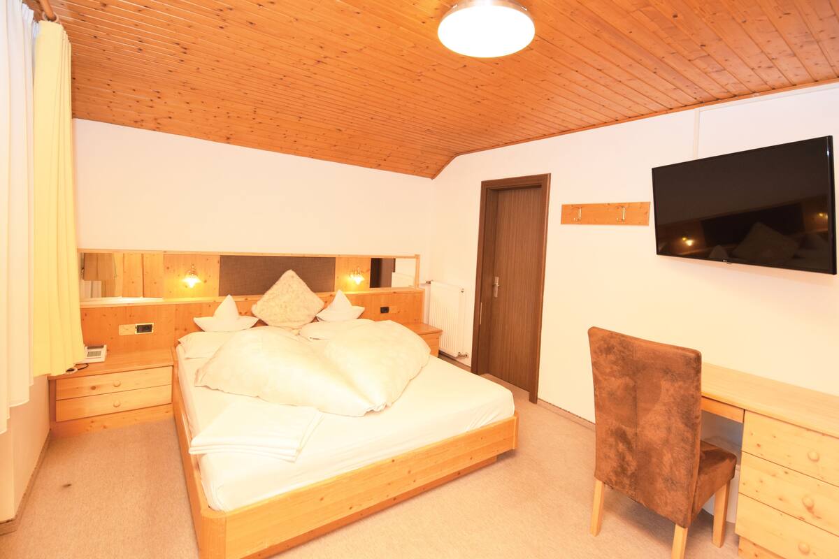 A bedroom with a wooden bed, bedside tables, a chair, a desk, and a TV on the wall.