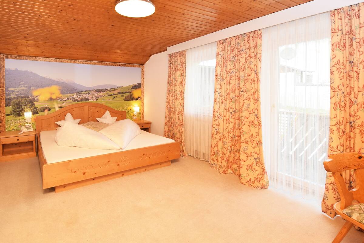A wooden-framed bed with white bedding in a room with wooden ceilings and large windows with floral curtains.