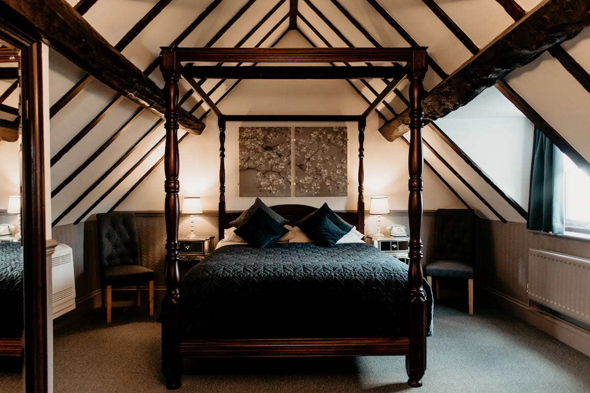 A four-poster bed in a room with sloped ceilings and exposed beams. There are two nightstands with lamps and a large painting on the wall.