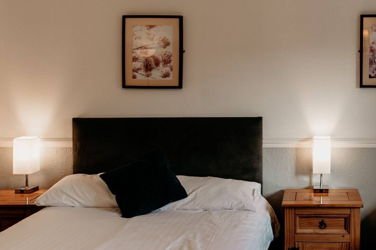 A neatly made bed with a black pillow, flanked by two bedside tables with lamps, in a room with a plain wall and a framed picture above the bed.
