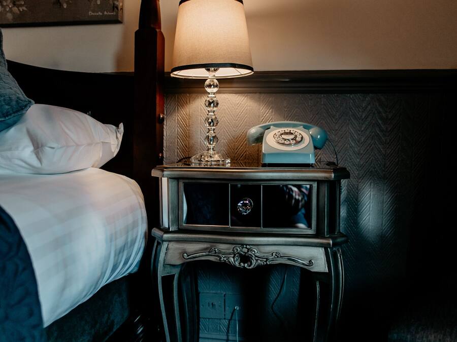 Un lit bien fait, avec du linge de lit blanc, une table de chevet avec une lampe et un téléphone d’époque.