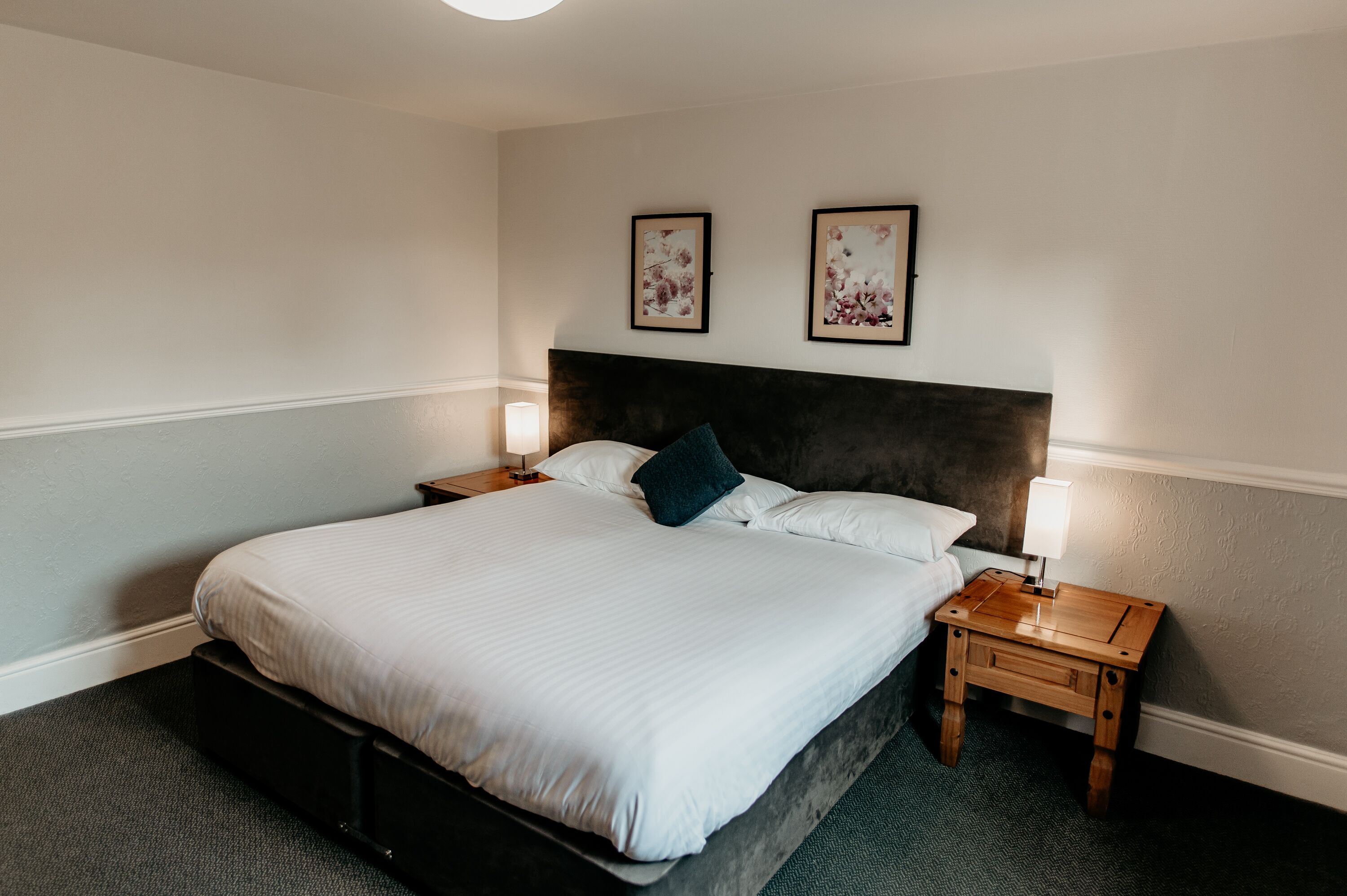 A neatly made bed with a dark headboard, two bedside lamps, and two framed floral artworks on the wall.