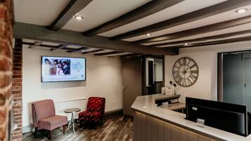 Un área de recepción con techo de madera, un reloj, un televisor mostrando un evento de boda, una silla roja estampada y una pequeña mesa.