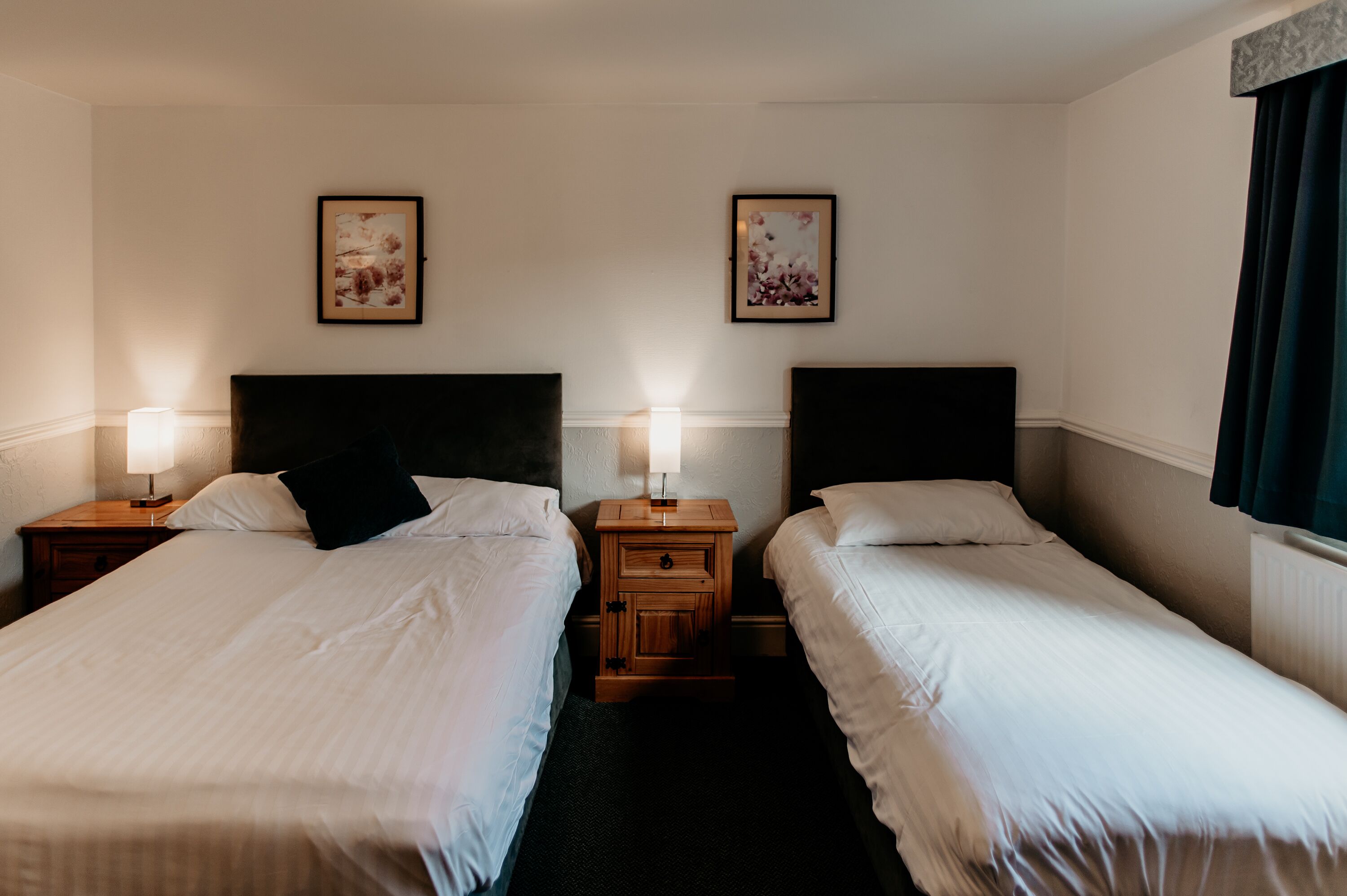A hotel room with two single beds, a wooden nightstand with a lamp, and two framed floral pictures on the wall.